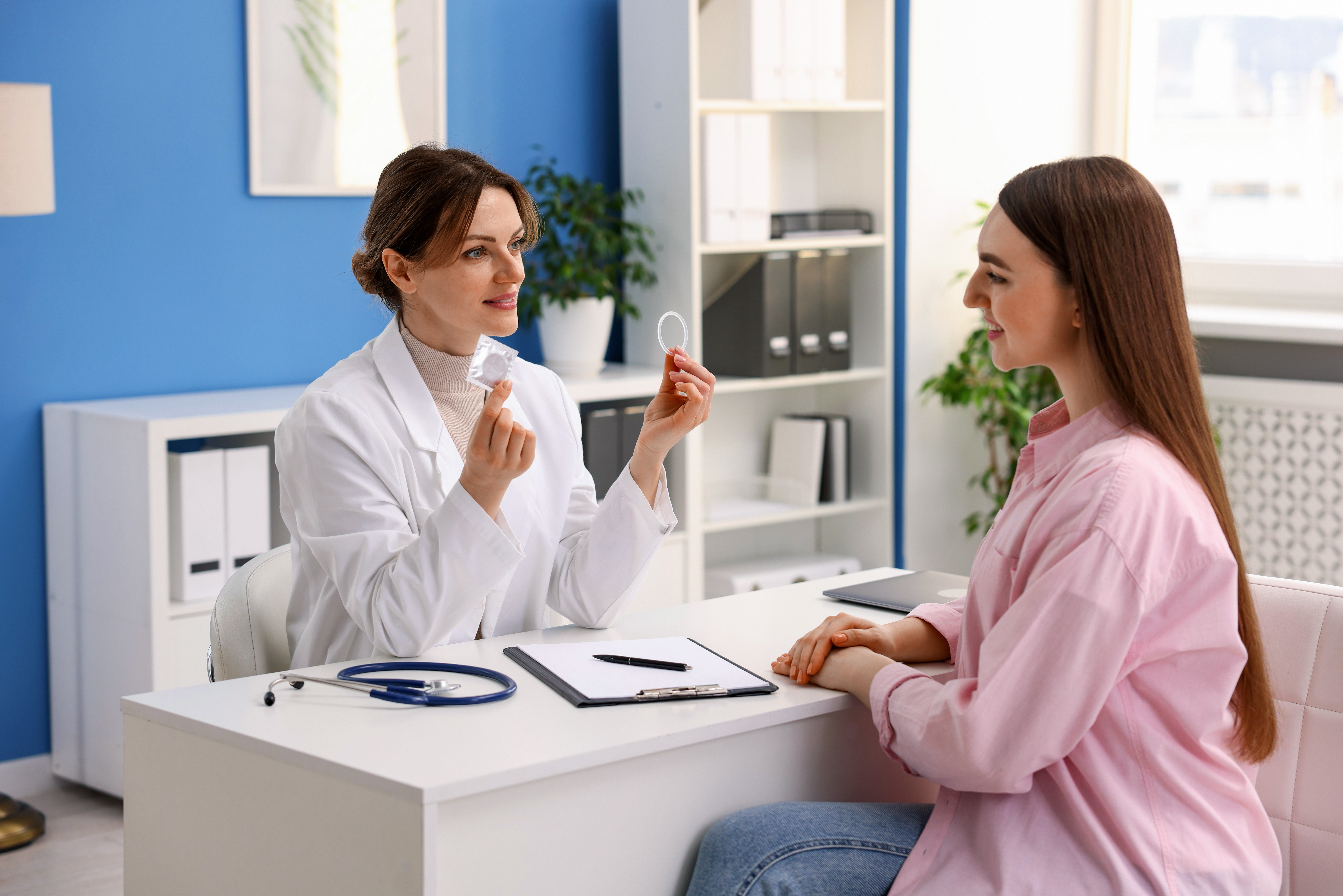 A female doctor sitting at a desk showing a condom and a vaginal ring to a female patient during a medical consultation.