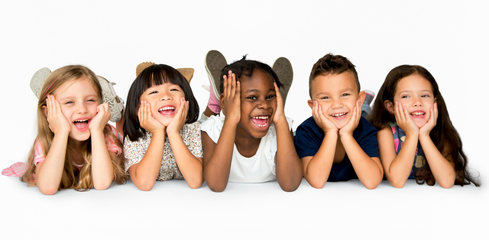 Five diverse young children lying on their stomachs in a row, resting their chins in their hands and smiling happily at the camera.